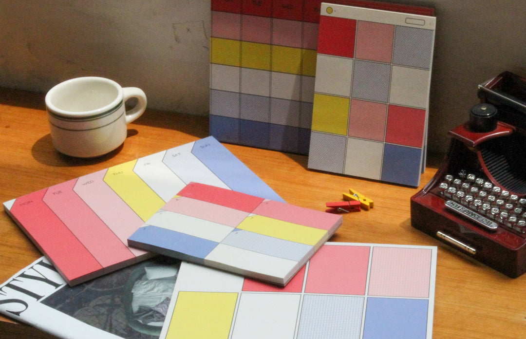 A set of colorful square sticky notes or cards with grid patterns in pink, yellow, blue, red, and white, arranged on a wooden desk alongside a white mug, a vintage red typewriter, and a magazine.