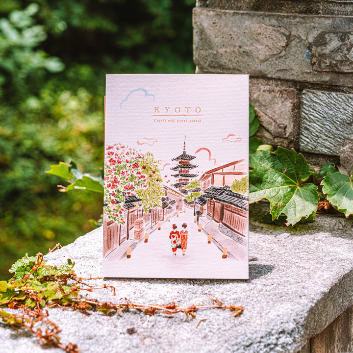A vertical art journal featuring a watercolor illustration of a Kyoto street with a pagoda and two children in kimonos, displayed on a stone ledge with green ivy and a stone wall background.