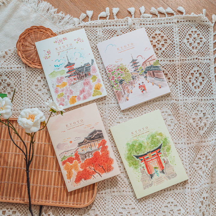 Four illustrated journals with Kyoto-themed watercolor designs are arranged on a cream lace textile over a bamboo tray, with white flowers and a small woven coaster nearby.