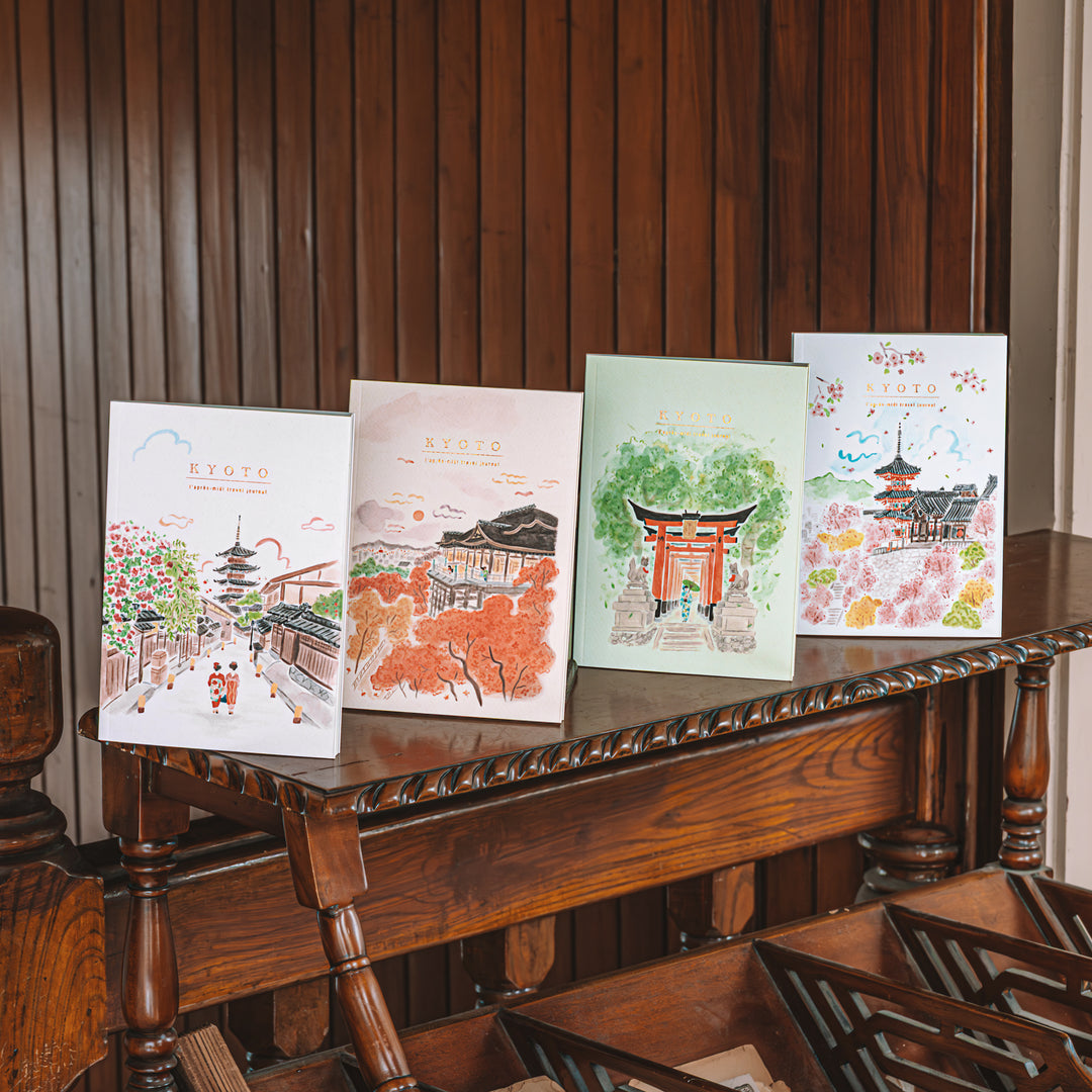 Four illustrated greeting journals with Japanese scenes are displayed upright on a carved wooden table against a dark wood-paneled wall.