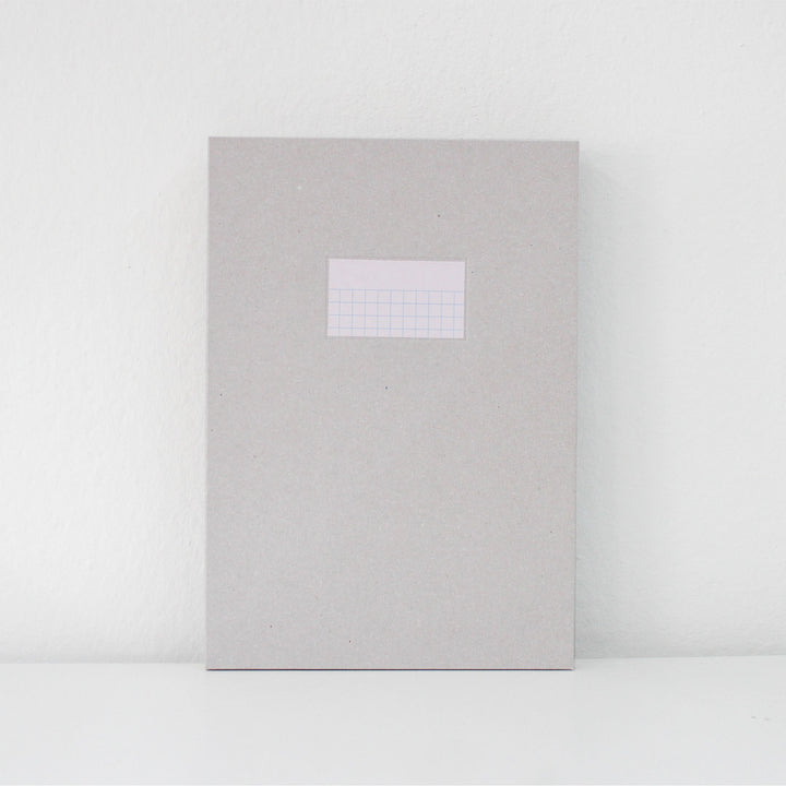 A light gray textured paper notebook or journal with a small white grid label on the front, standing upright against a white wall on a white surface.
