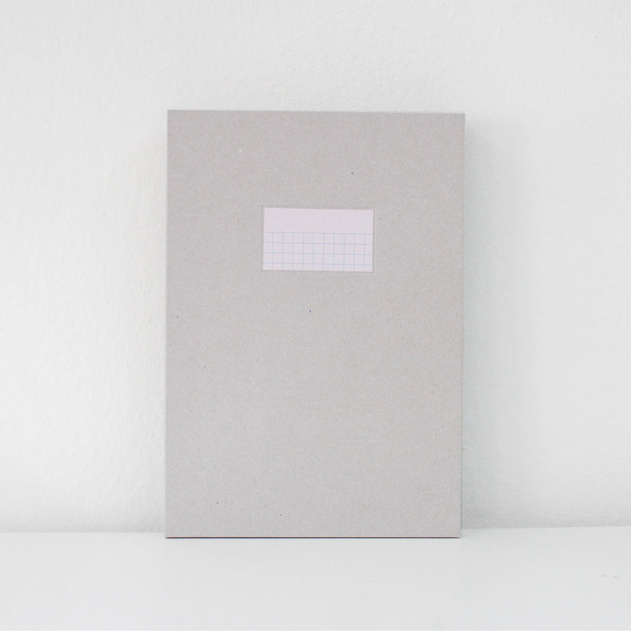 A light gray textured paper notebook or journal with a small white grid label on the front, standing upright against a white wall on a white surface.