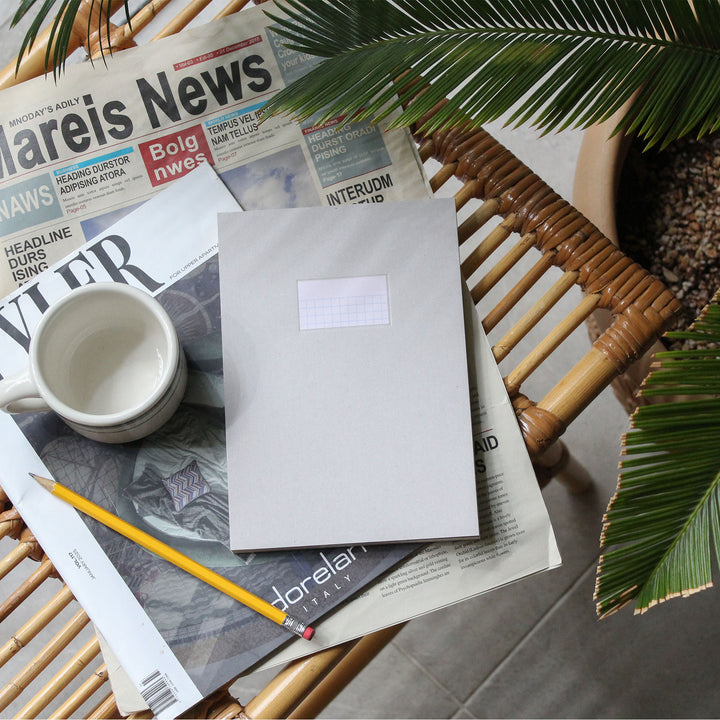 A plain light gray envelope with a small blank white label sits on top of stacked newspapers on a woven rattan chair, with a white mug, yellow pencil, and green plant leaves around it.