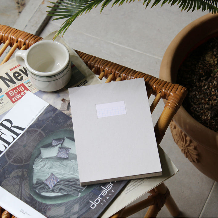 A light gray fabric-covered notebook or journal with a blank white label on the front, styled on top of magazines on a wicker chair next to a white ceramic mug and a potted plant.