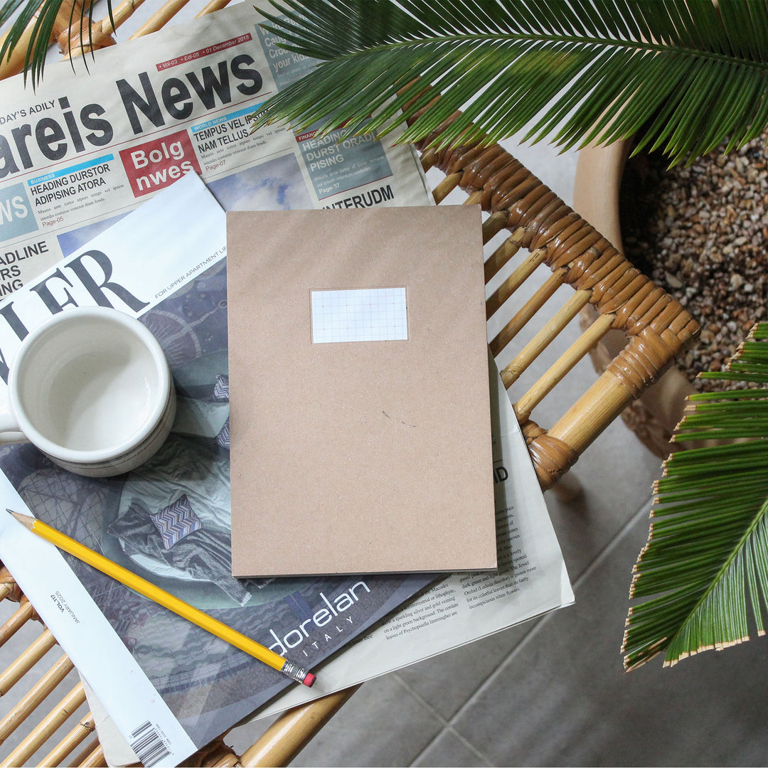 A kraft paper notebook with a blank white label on the front sits on a wicker chair, styled with a white mug, a yellow pencil, and various printed newspapers, with green palm leaves framing the scene.