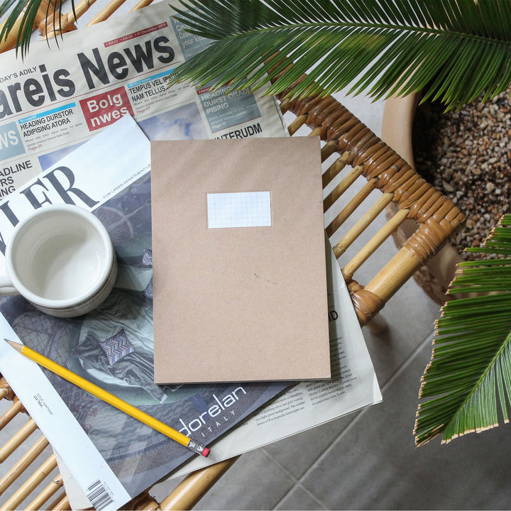 A kraft paper notebook with a blank white label on the front sits on a wicker chair, styled with a white mug, a yellow pencil, and various printed newspapers, with green palm leaves framing the scene.