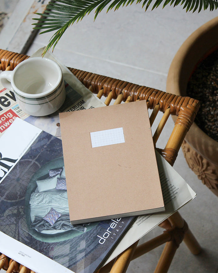 A kraft paper notebook with a small white label sits on top of magazines on a woven rattan chair, next to a white ceramic mug and a potted plant.