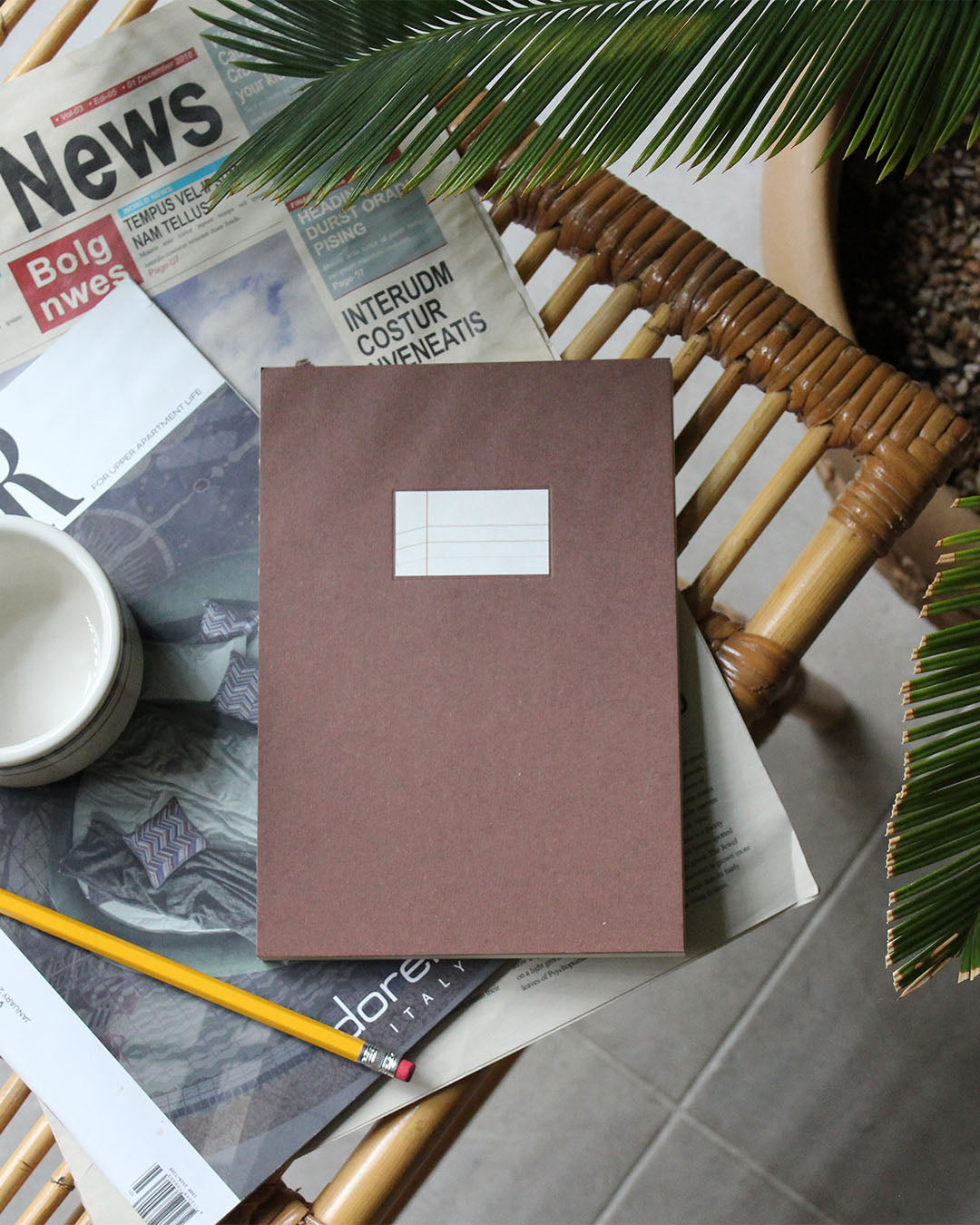 A brown paper notebook with a blank white label on the front is arranged on a wicker chair in a styled flat-lay setting with magazines, a pencil, a small cup, and plant leaves.