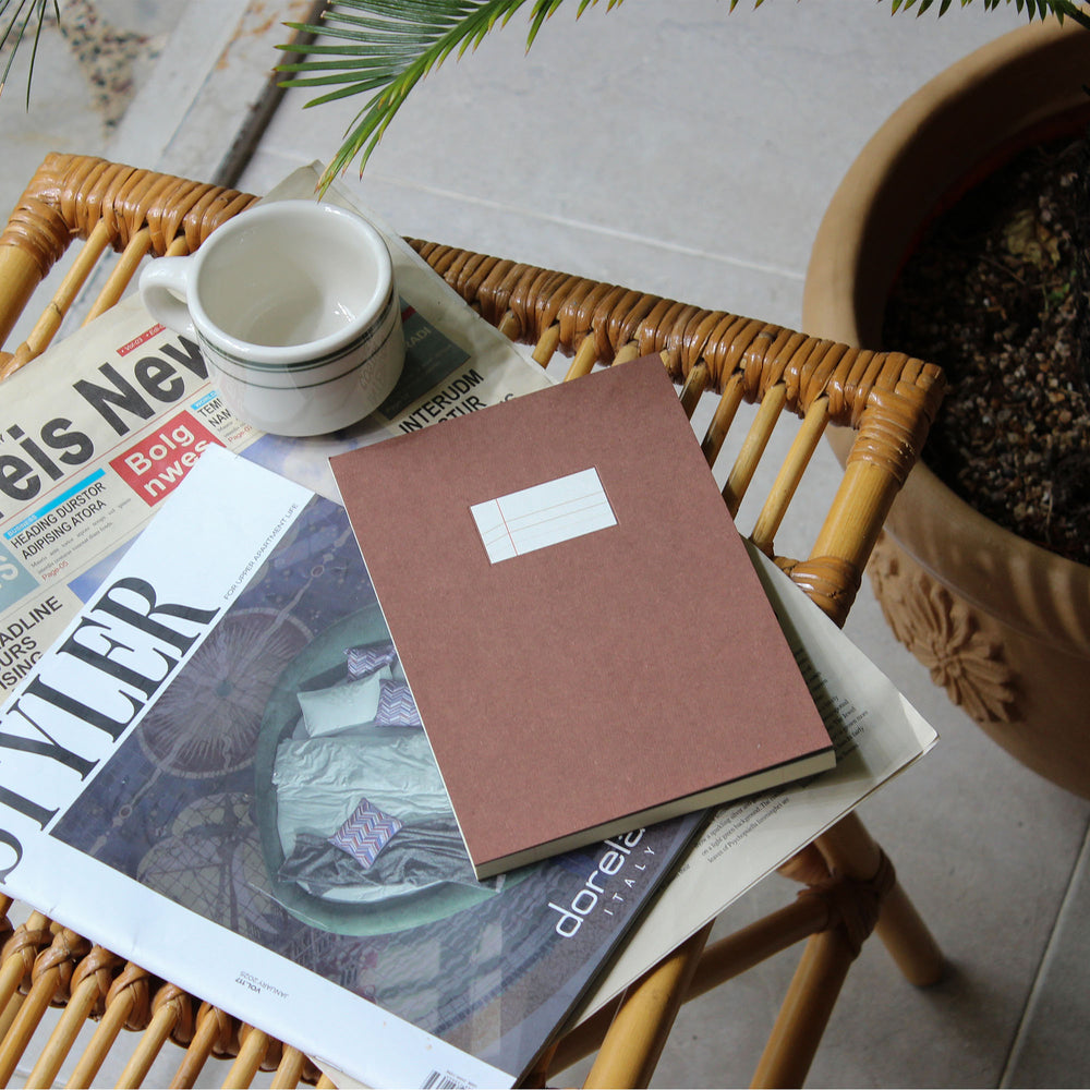 A brown hardcover notebook with a blank white label sits on top of magazines on a woven rattan chair, next to a white ceramic mug and a potted plant.